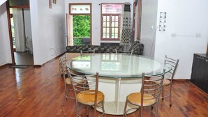 A spacious room with a round glass table surrounded by metal chairs, large windows with curtains, and a view of greenery outside. - Srivari Studio Apartment (Kodaikanal)