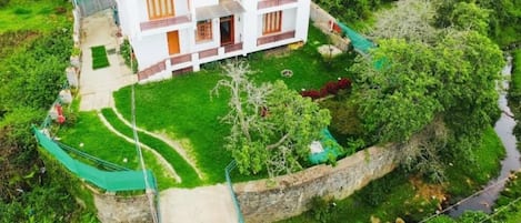 Un appartement-studio moderne à deux étages, doté d’un toit plat, entouré d’une végétation luxuriante et traversé par un petit ruisseau.