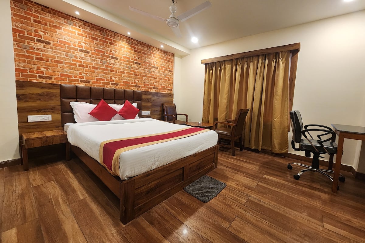 Habitación de hotel con pared de ladrillo, cabecero de madera, cama con ropa blanca y almohadas rojas, un escritorio con silla y un ventilador de techo.
