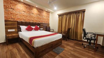 Une chambre d’hôtel avec un mur en briques, une tête de lit en bois, un lit avec une literie blanche et des oreillers rouges, un bureau avec une chaise et un ventilateur de plafond.