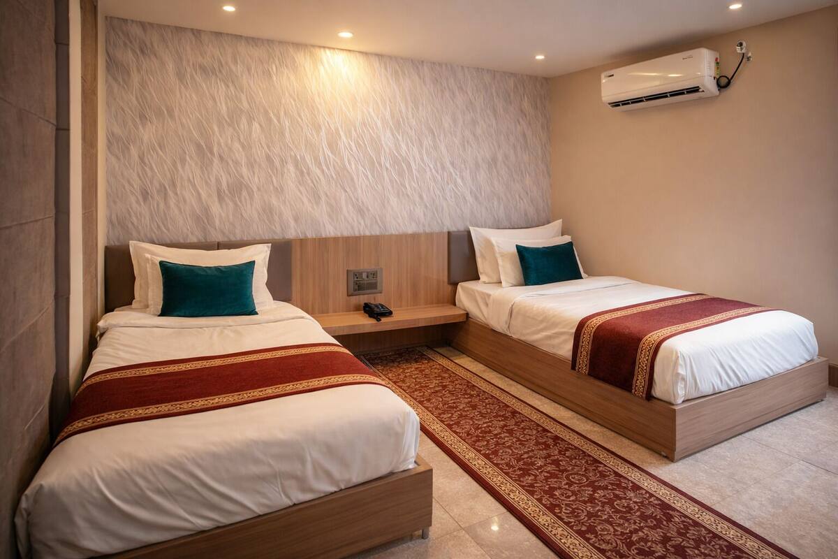 A hotel room with two single beds, a wooden headboard, a red and gold patterned rug, and an air conditioning unit on the wall.