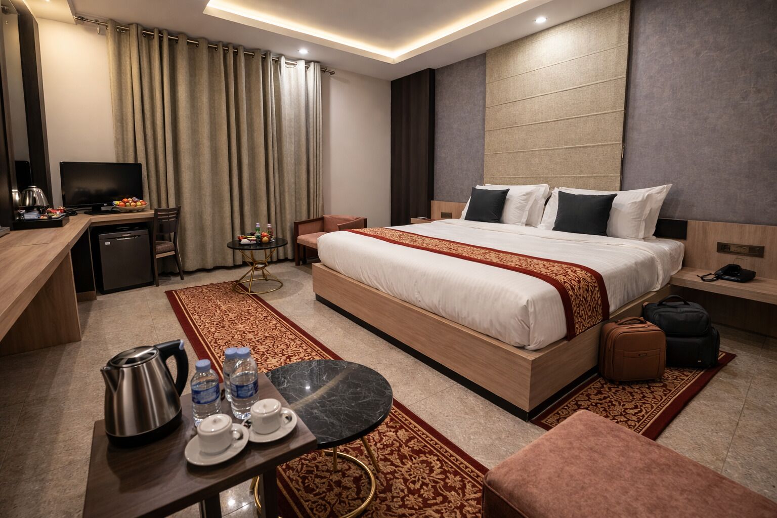 A hotel room with a large bed, a desk with a kettle and cups, a TV, and a small table with fruit and a bottle.