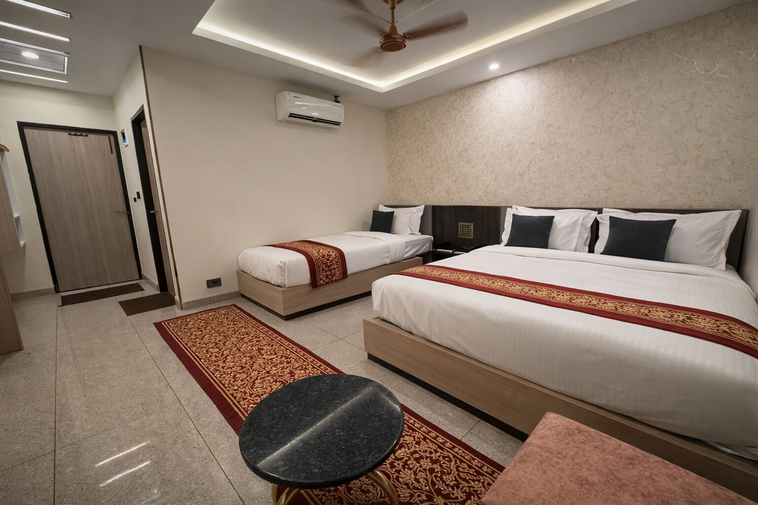 A hotel room with two beds, a ceiling fan, an air conditioning unit, a small round table, and a red and gold patterned rug.