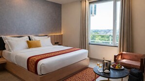 A hotel room with a large bed, a bedside table, a small round table with drinks and fruit, a chair, and a view of the city through the window. - OPO Horizon Surbhi Grant,Khatu Shyam (Danta Ramgarh)