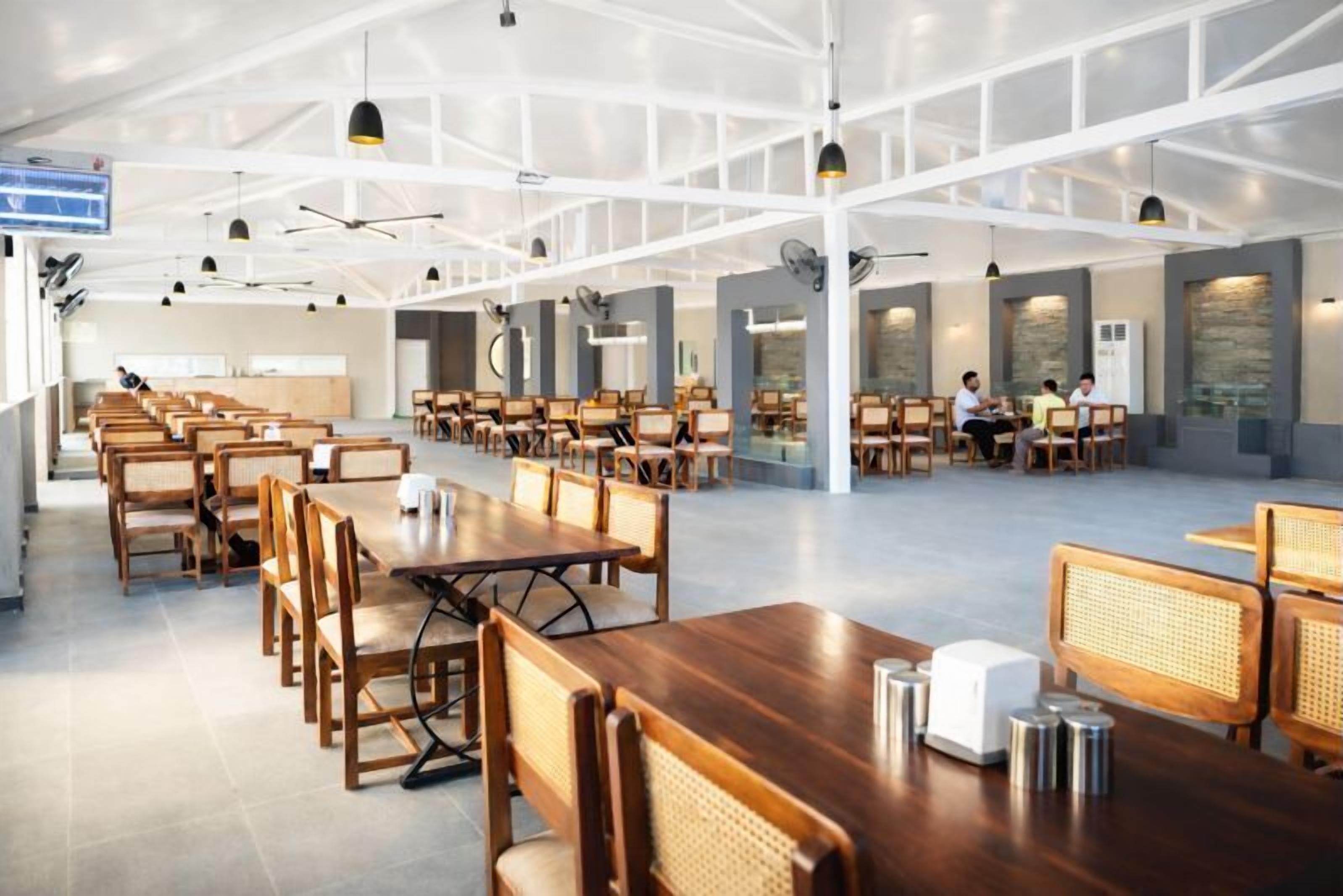 Un'ampia sala da pranzo con tavoli e sedie in legno, travi a soffitto bianche e illuminazione a sospensione.