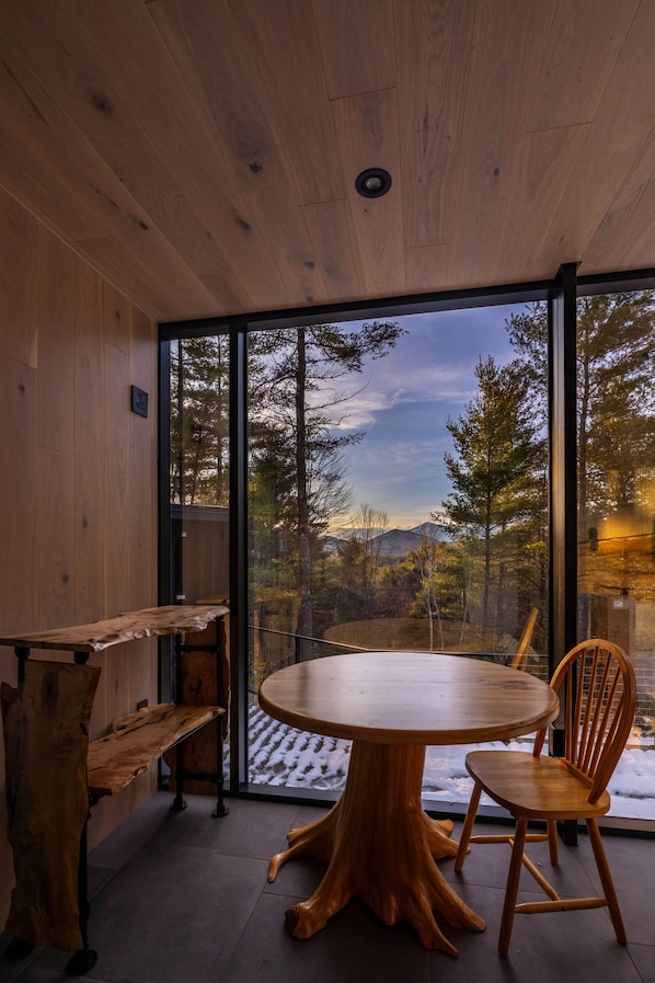 A wooden interior with a round table and chairs, a bench, and a large glass door leading to a forested outdoor area.