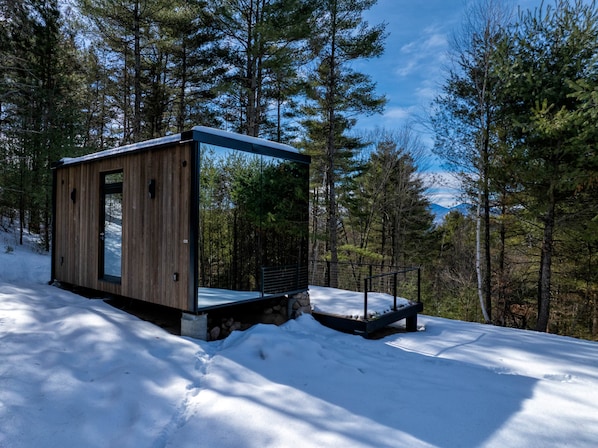 Une cabane en bois moderne, dotée d’une porte en verre, entourée de neige et d’arbres.