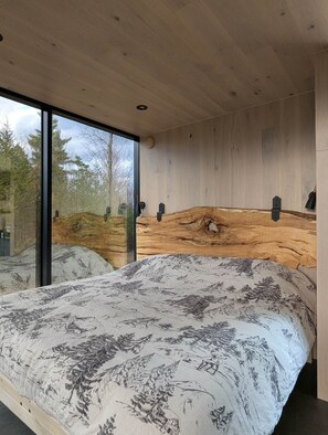 A modern bedroom with a large bed, wooden headboard, and a view of trees through large windows.
