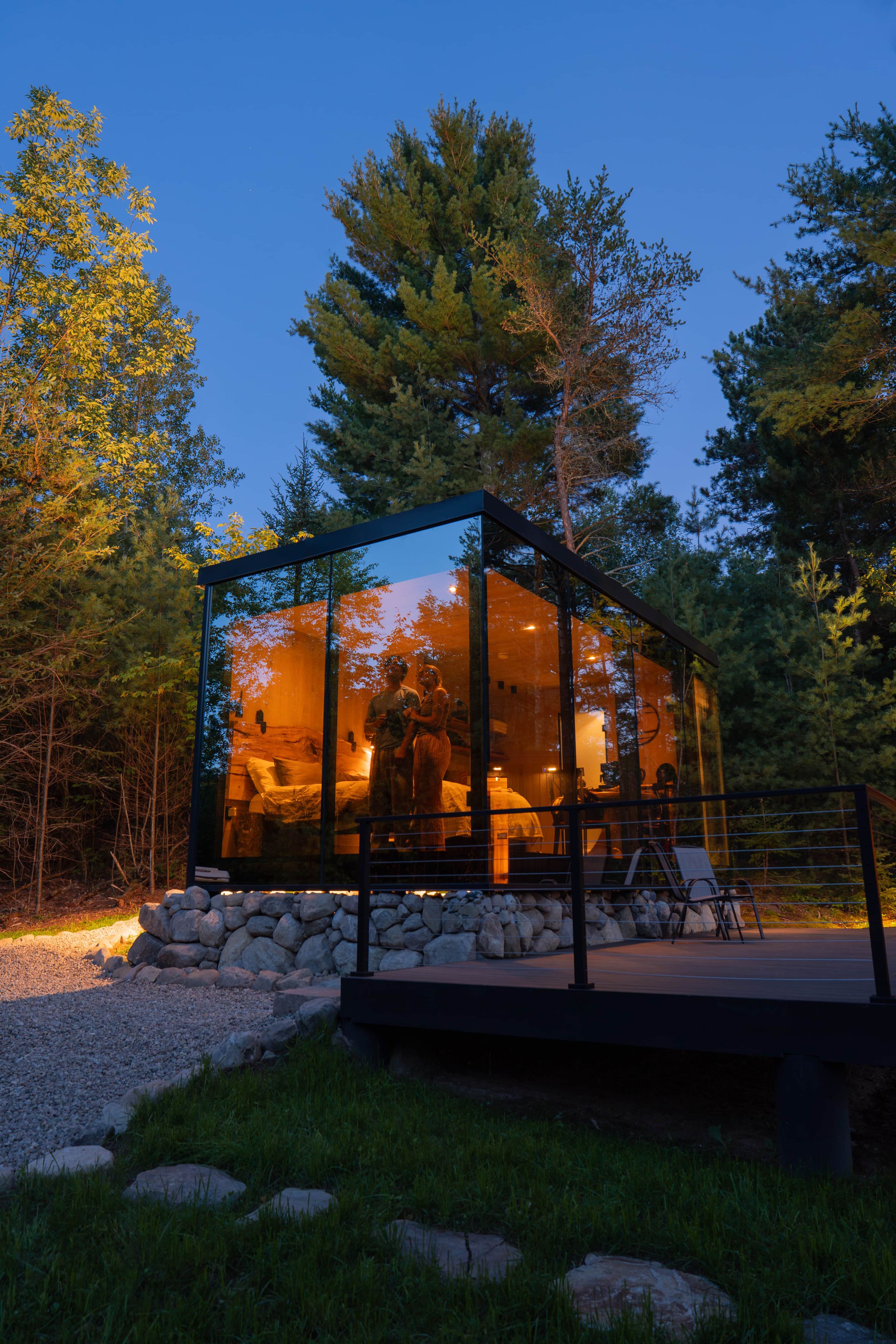 A modern glass structure with a stone base, surrounded by a natural landscape with trees and a gravel path.