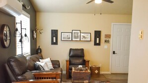 A living room with a leather couch, wooden coffee table, and a leather chair. There is a ceiling fan, a rug, and framed pictures on the wall. - Cowboy Condo in Charming Shelby (Shelby)