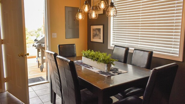 A dining area with a wooden table, six chairs, hanging light fixtures, a potted plant, and a window with blinds. - Cowboy Condo in Charming Shelby (Shelby)