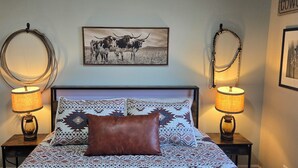 A bed with patterned bedding, a brown pillow, and two wall-mounted lamps. - Cowboy Condo in Charming Shelby (Shelby)