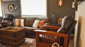 A cozy living room with a leather sofa, a wooden armchair, a vintage suitcase, and a rustic rug. - Cowboy Condo in Charming Shelby (Shelby)