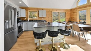 A modern kitchen with a central island, bar stools, and a dining area with a view of the outdoors. - Fox Run Log Cabin~ LUXE~Asheville~Dutch Cove (Canton)