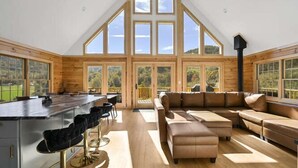 A spacious room with a high ceiling, large windows, a central seating area, and a kitchen counter. - Fox Run Log Cabin~ LUXE~Asheville~Dutch Cove (Canton)