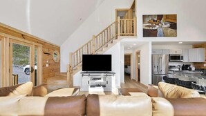 A spacious living area with a large leather sofa, a flat-screen TV, wooden flooring, a staircase, and a kitchen in the background. - Fox Run Log Cabin~ LUXE~Asheville~Dutch Cove (Canton)