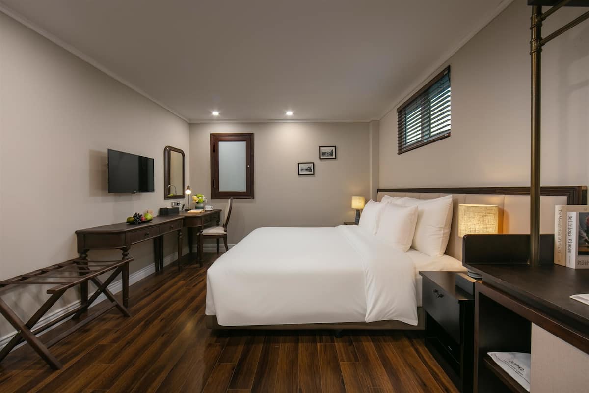 Une chambre d’hôtel avec un grand lit, un bureau avec une chaise, une télévision et une fenêtre avec des stores.