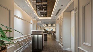 Pasillo de hotel con mostrador de recepción central, un área de comedor al fondo y un techo con conductos a la vista.