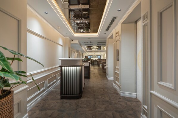 Un couloir d’hôtel avec un comptoir de réception central, une salle à manger à l’arrière-plan et un plafond laissant apparaître les conduits.