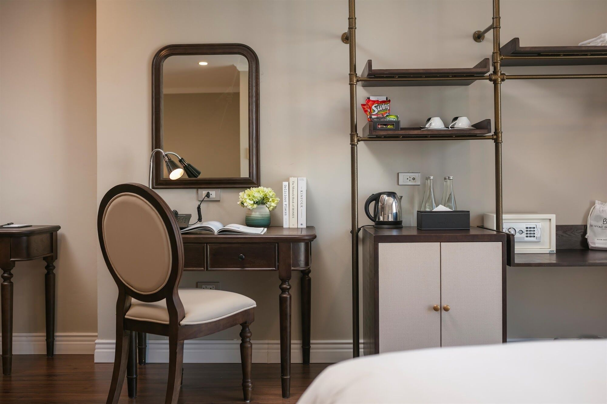 Een hotelkamer met een houten bureau, een stoel, een bed, een spiegel, een plank met toiletartikelen en een waterkoker.
