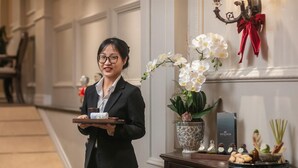 Un miembro del personal de hotel lleva una bandeja con una toalla y una bebida, de pie junto a un jarrón decorativo con flores blancas y una estación de bienestar con varios artículos.