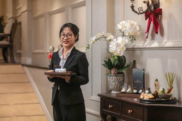 Een personeelslid van een hotel draagt een dienblad met een handdoek en een drankje, staand naast een decoratieve vaas met witte bloemen en een wellnessstation met diverse artikelen.