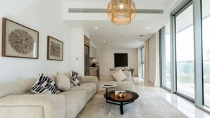 A modern living room with a beige sofa, a coffee table, and a dining area with a chandelier. - Beach Isle Residences- Emaar Beachfront (Dubai)