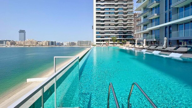 A swimming pool with clear blue water, surrounded by modern buildings and a beach in the background. - Beach Isle Residences- Emaar Beachfront (Dubai)