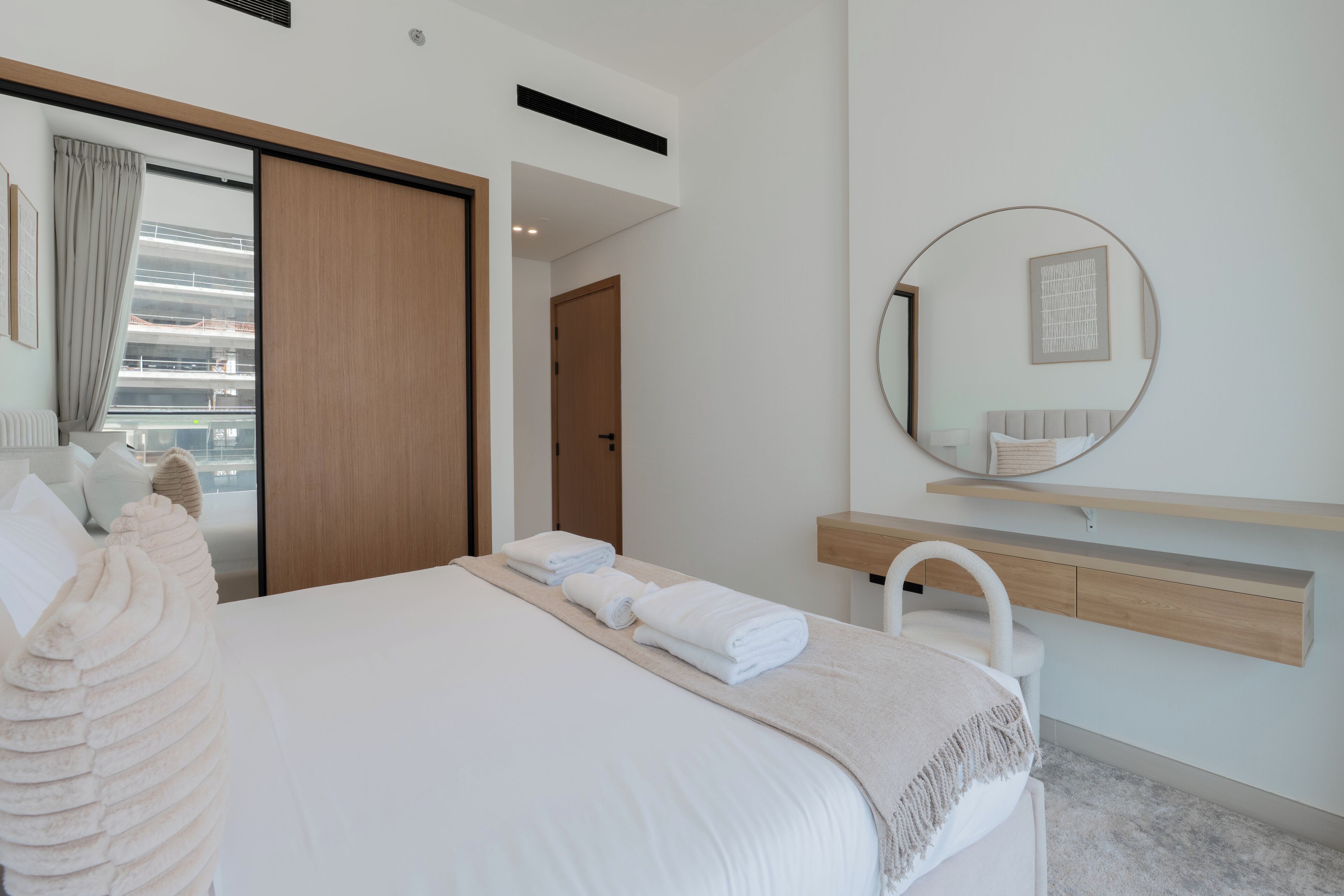 Una habitación de hotel moderna con una cama grande, un armario de madera, un espejo redondo y vistas a un edificio desde la ventana.