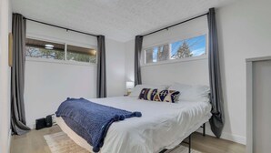 A modern bedroom with a large bed, grey curtains, a window, and a view of trees. - The Penick Getaway Su (Reynoldsburg)