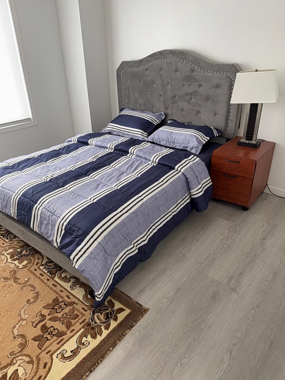 A neatly made bed with striped bedding, a grey upholstered headboard, a bedside table with a lamp, and a patterned rug on a wooden floor.