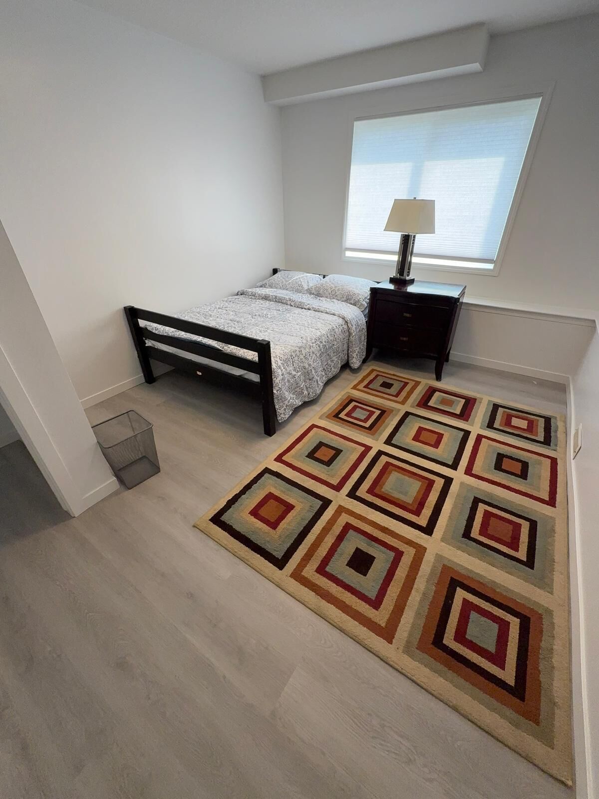 A bedroom with a bed, a nightstand, a lamp, a window, and a patterned rug.