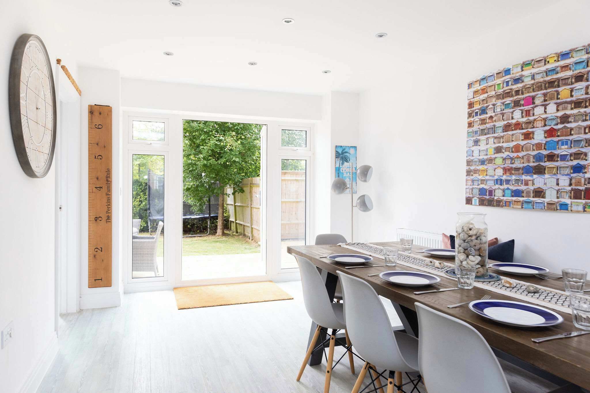A dining area with a wooden table set for four, white chairs, a large wall clock, a family rule measuring tape, and a colorful wall art piece.
