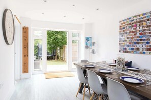 A dining area with a wooden table set for four, white chairs, a large wall clock, a family rule measuring tape, and a colorful wall art piece. - Modern Family Retreat Nr Beaches and Marina (Birdham)
