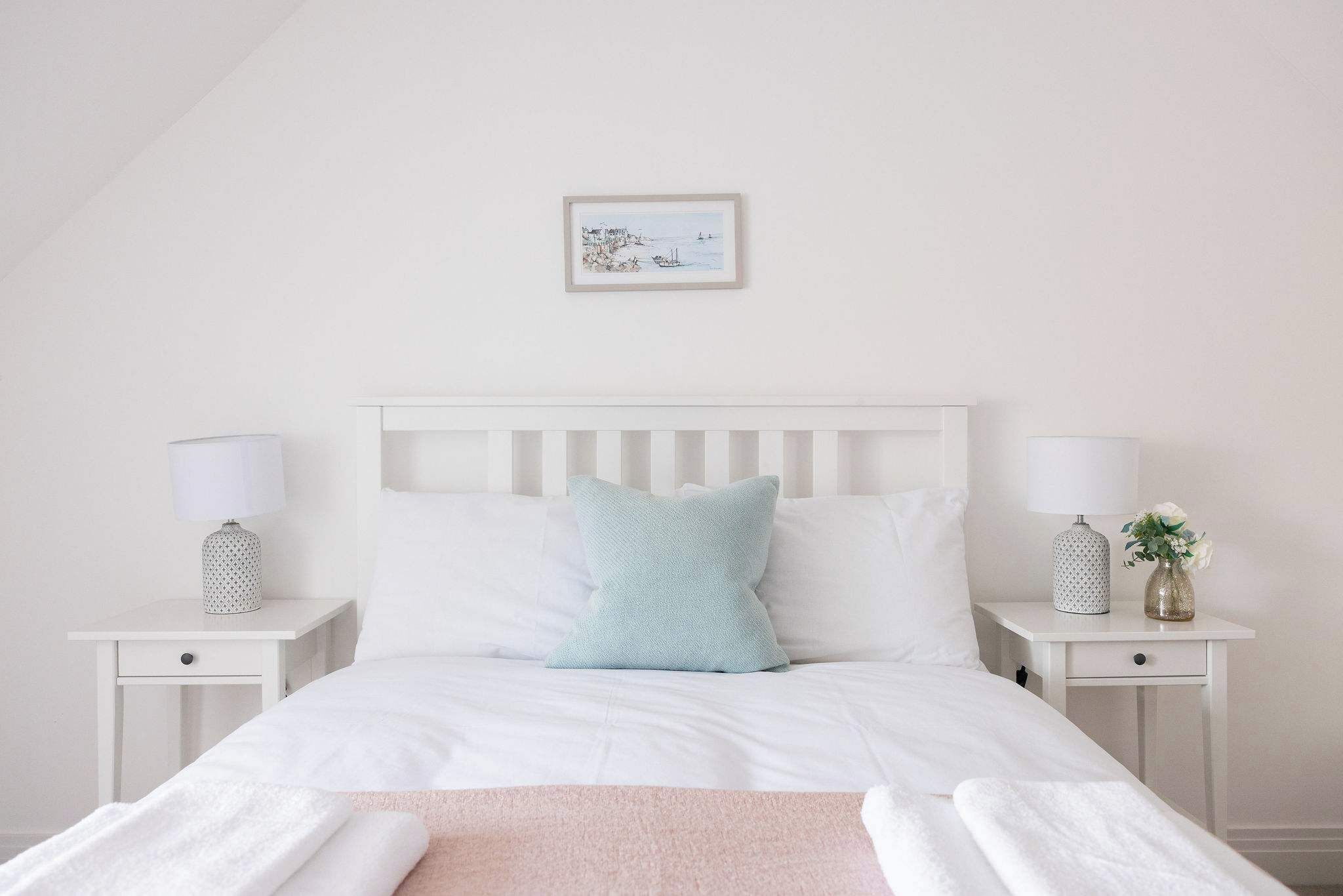 A neatly arranged bedroom with a white bed, two bedside tables with lamps, a small framed picture on the wall, and a pink throw blanket.