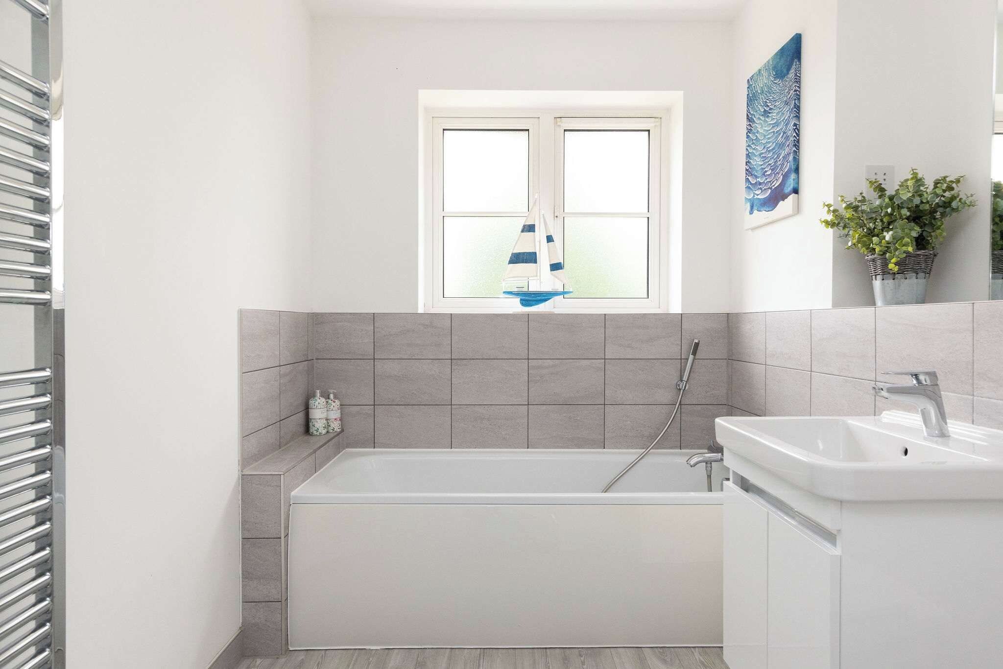 A modern bathroom with a bathtub, a window with a sailboat decoration, and a potted plant.