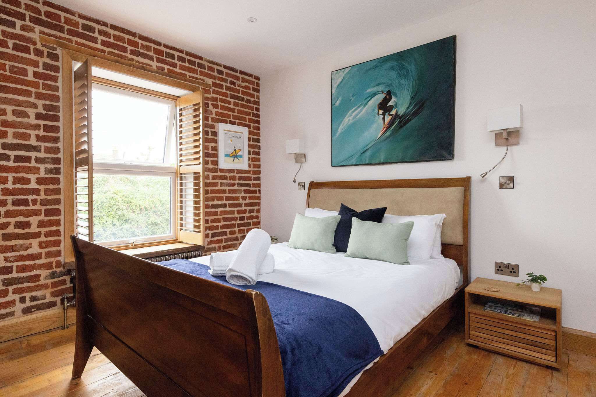 A bedroom with a wooden bed, a brick wall, a window with blinds, a painting of a surfer, and a nightstand with a plant.