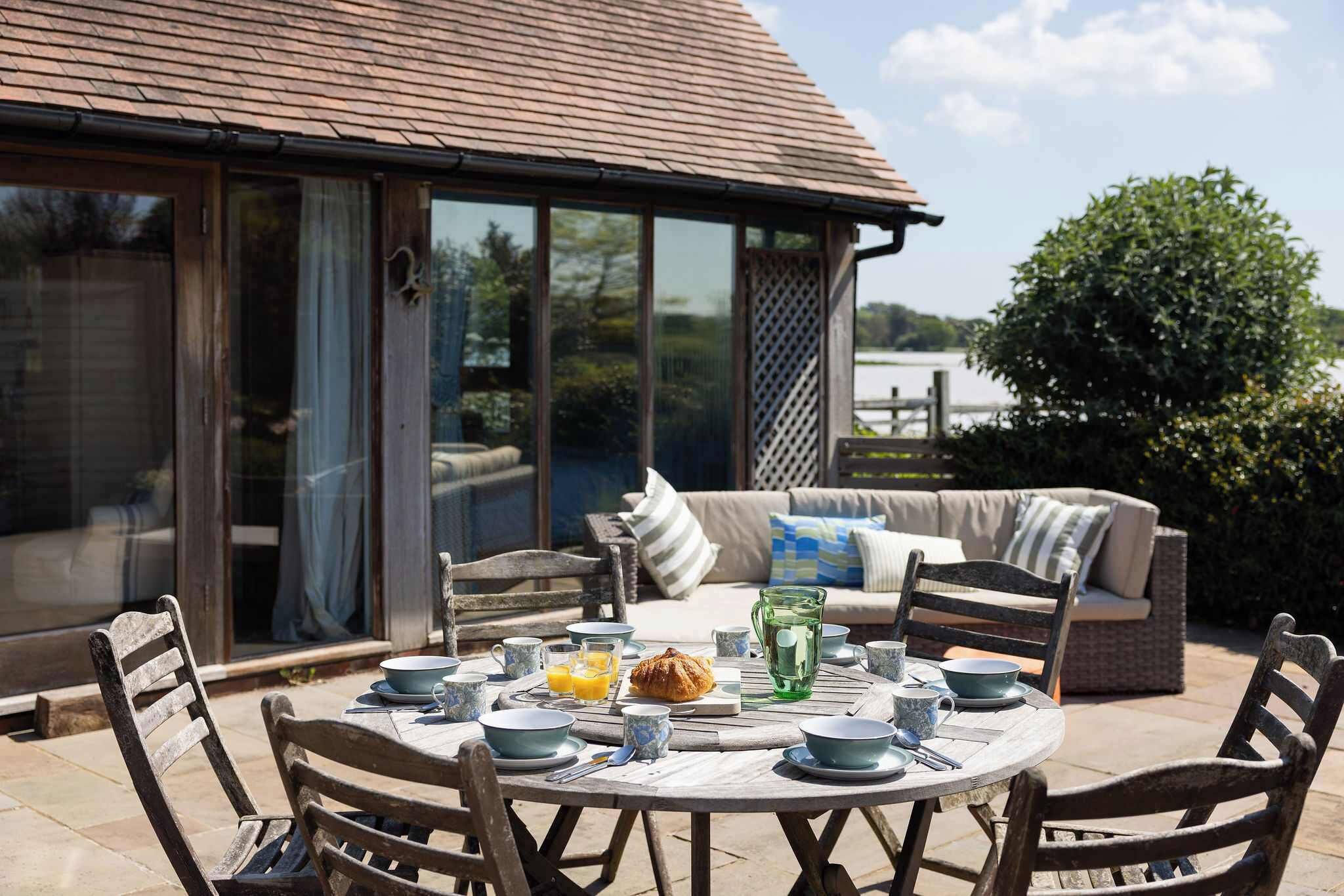 Une table ronde dressée avec des aliments pour le petit-déjeuner, entourée de chaises en osier, avec vue sur un lac et un espace de détente extérieur abrité.