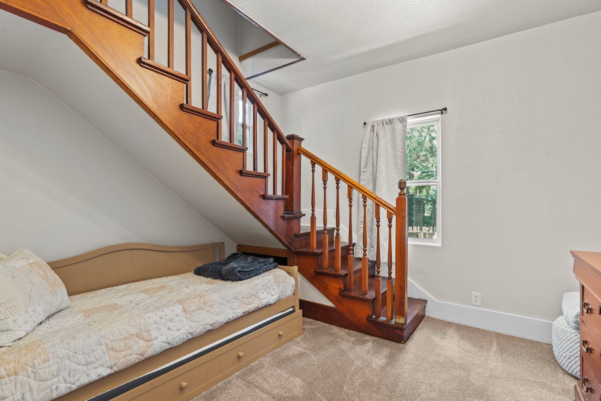 A bedroom with a wooden staircase, a bed, a dresser, and a window with a view of greenery.