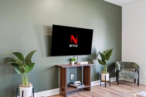 Una sala de estar con televisor de pantalla plana montado en la pared, una consola de madera, dos plantas en macetas, un sillón verde y una estantería.