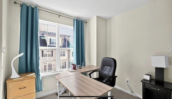 Habitación de hotel con un escritorio de madera, una silla de oficina negra, una lámpara blanca y una ventana con cortinas color turquesa.