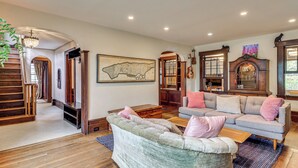 A spacious living room with a large green sofa, a grey sofa, and a wooden coffee table. There is a staircase leading to an upper level, a large map on the wall, and a wooden cabinet with a cat figurine on top. - Easy Train Access to NYC! Elegant Family Retreat (West Orange)