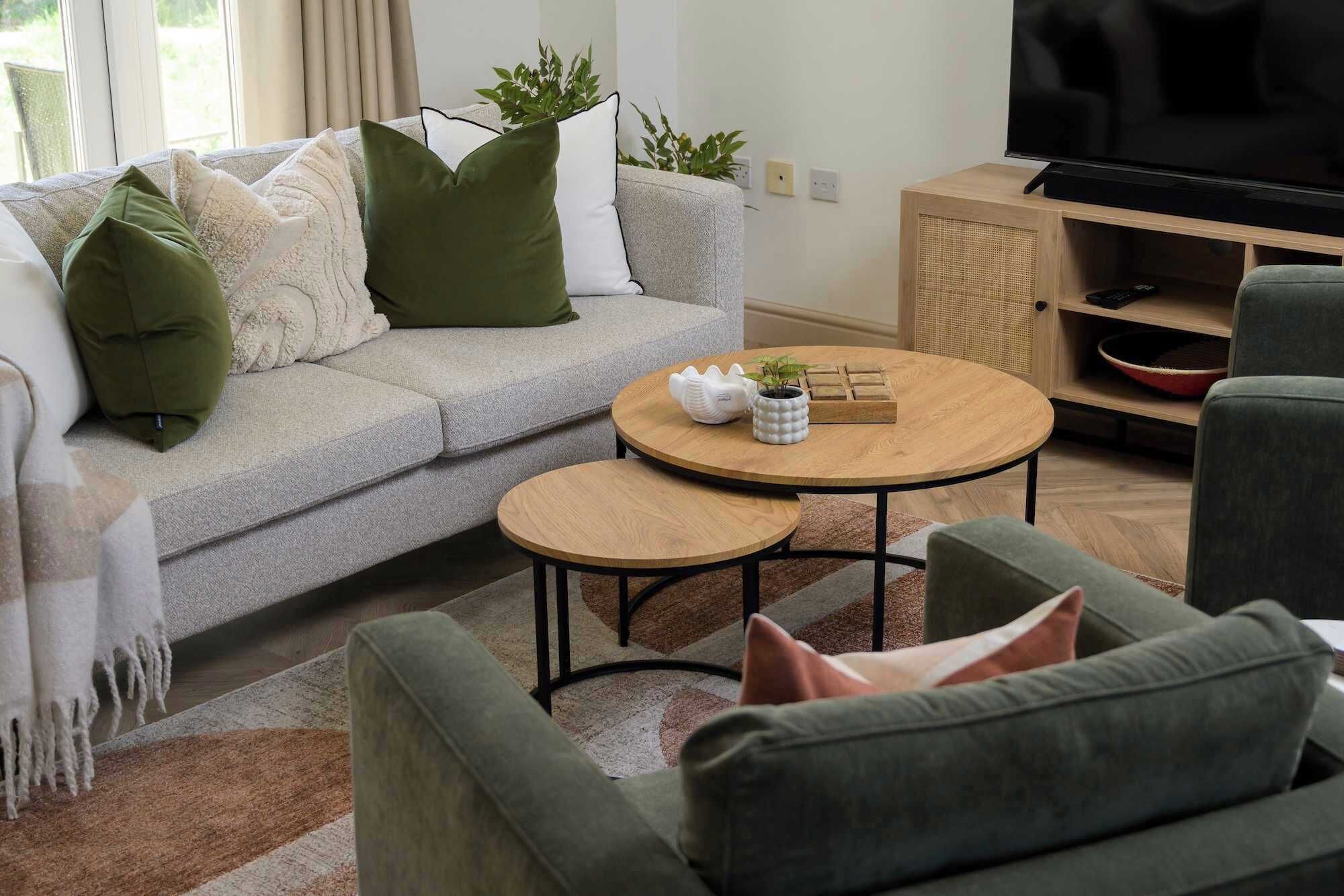Una sala de estar con un sofá gris, dos mesas redondas de madera y un televisor sobre un mueble de madera.