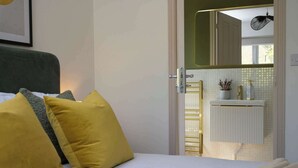 Quarto com cama, almofadas, mesa de cabeceira, espelho e vista para um banheiro com radiador e planta.