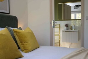 Et soveværelse med seng, puder, et natbord, et spejl og udsigt til et badeværelse med en radiator og en plante.