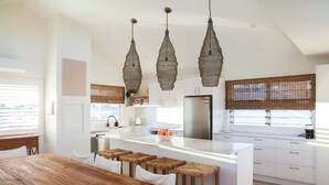 A modern kitchen with a central island, white cabinetry, and a wooden dining table. - The Nest - Oceanstays (Yamba)