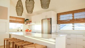 A modern kitchen with white cabinetry, a stainless steel refrigerator, wicker bar stools, and a large window with blinds. - The Nest - Oceanstays (Yamba)