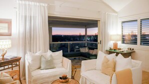 A spacious living area with a balcony, white furniture, and a view of the cityscape at dusk. - The Nest - Oceanstays (Yamba)