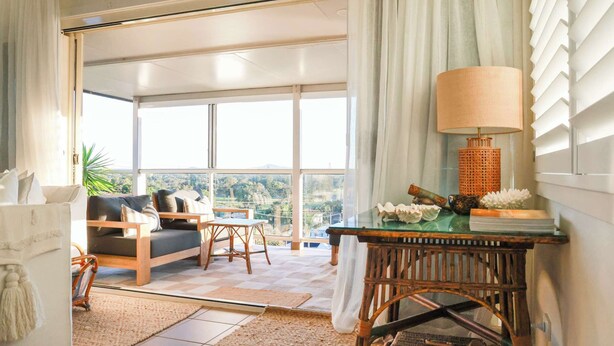 A spacious living area with a sofa, chairs, a table, and a view through large windows. - The Nest - Oceanstays (Yamba)