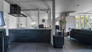 A modern kitchen with dark cabinetry, a central island, and a built-in stove. There is a large window providing natural light and a view of the outdoors. - GROUNDBLOOM YIG STAY (Furano)